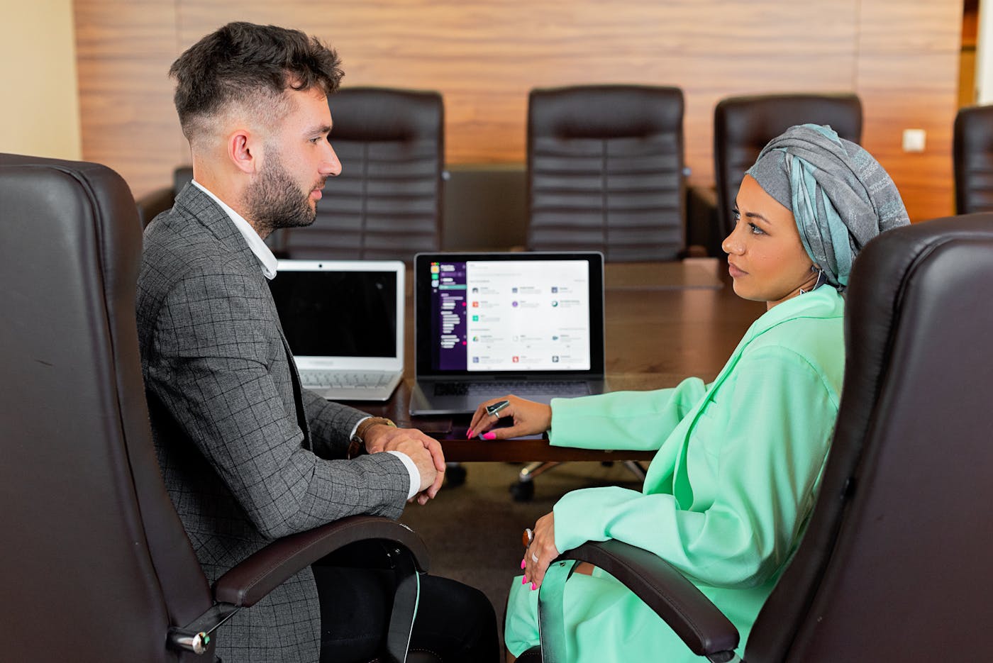 Two professionals discussing work across a boardroom table with laptops nearby.